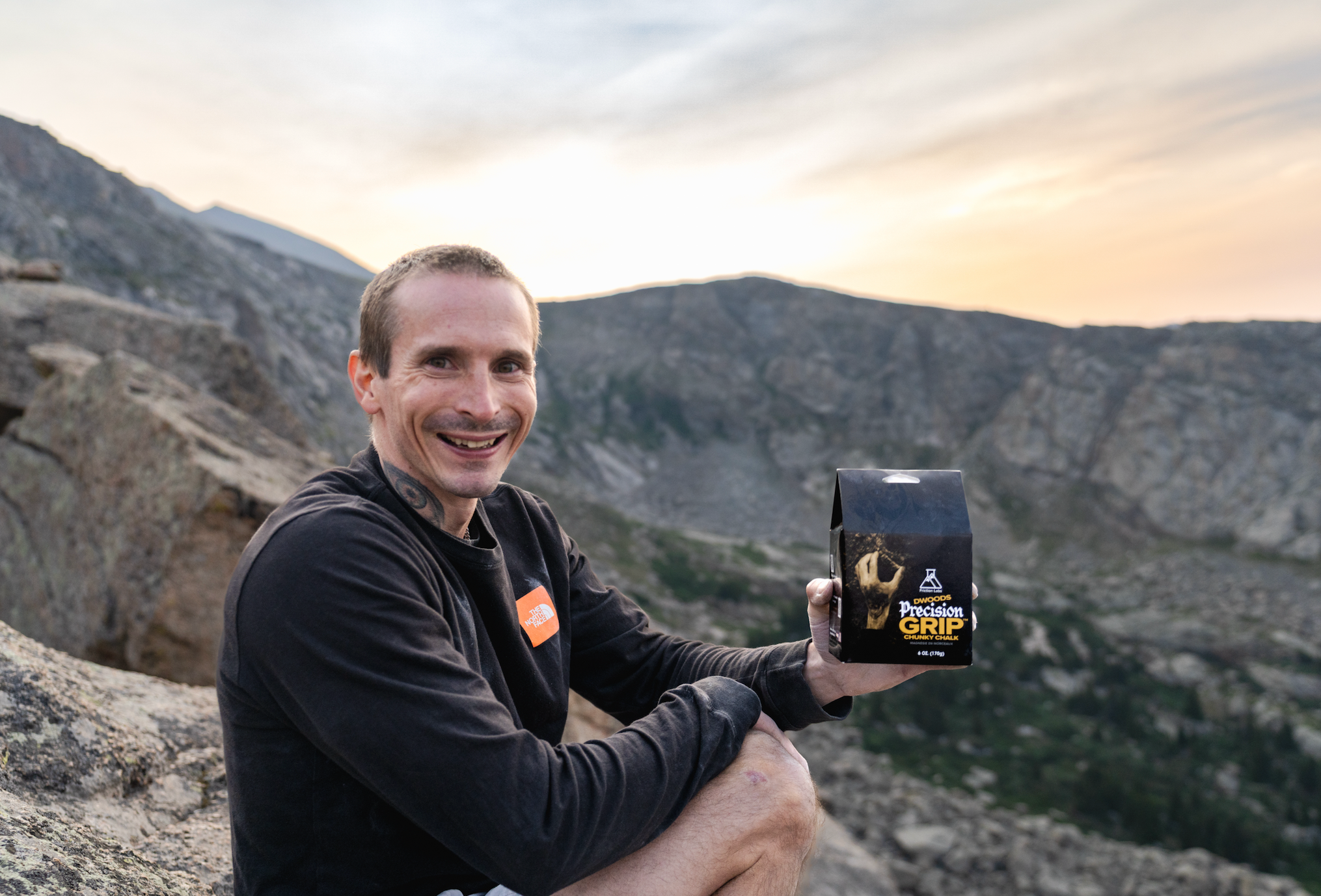 Pro Climber Daniel Woods in a black long-sleeve shirt sits outdoors on a rocky mountain slope at sunset, holding up a black bag of DWoods Precision Grip Chunky Loose Chalk. The background shows rugged mountains and a soft sky.